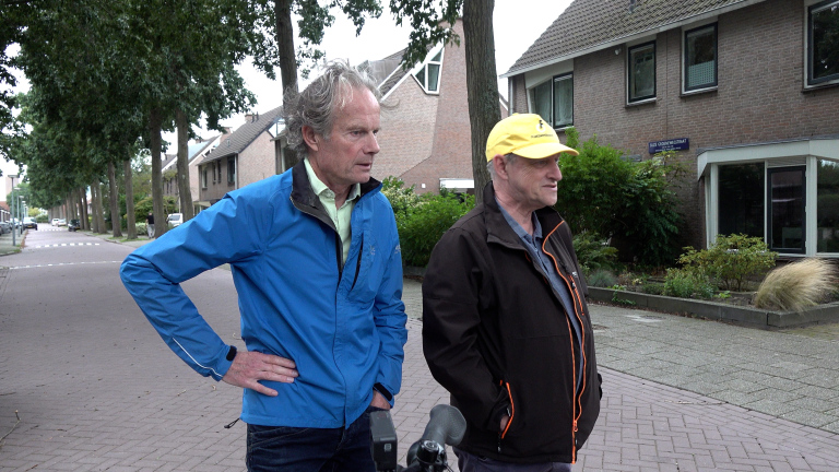 Twee mannen staan op een woonstraat; één in blauwe jas, de ander met gele pet, met microfoons op de voorgrond