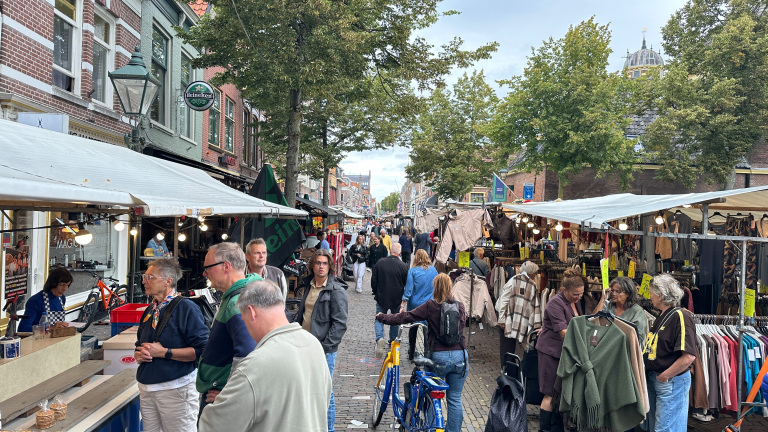 Drukke straatmarkt met kramen vol kleding en etenswaren, shoppers tussen bomen en oude gevels op een klinkerstraat