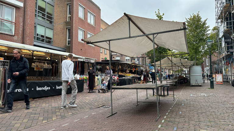 Straatmarkt met kramen en bezoekers, lege tafels onder luifels op een geplaveid plein tussen bakstenen gebouwen
