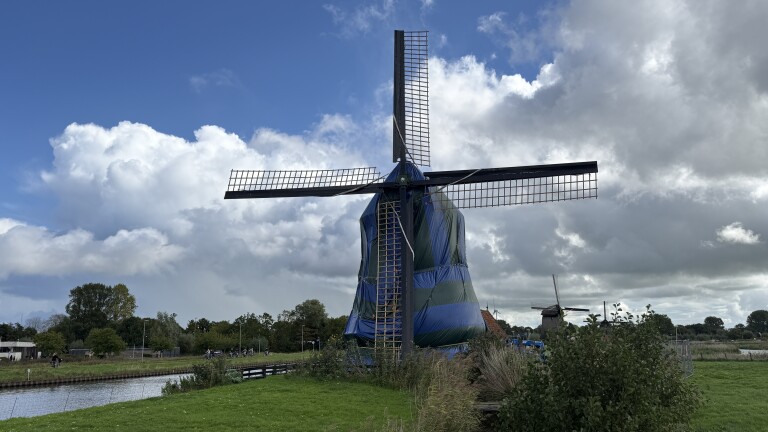 Windmolen in een groen-blauw zeilomhulsel langs een kanaal, met wieken tegen een wolkenlucht