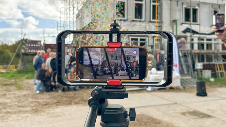 Smartphone op statief filmt een groep mensen voor een gebouw, met op het scherm een opname-timer en recordknop