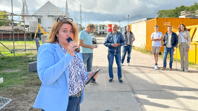 Vrouw spreekt in microfoon met telefoon in hand, met groep mensen op bouwterrein en oranje container op de achtergrond