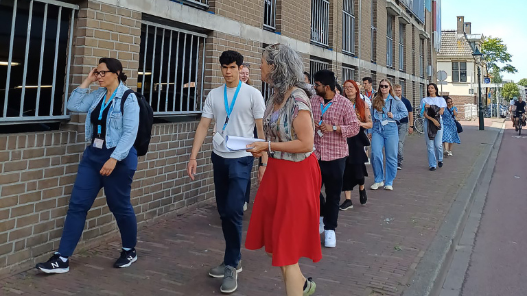 Groep mensen met blauwe naambadges loopt langs een bakstenen gebouw, geleid door een vrouw met grijs haar en rode rok