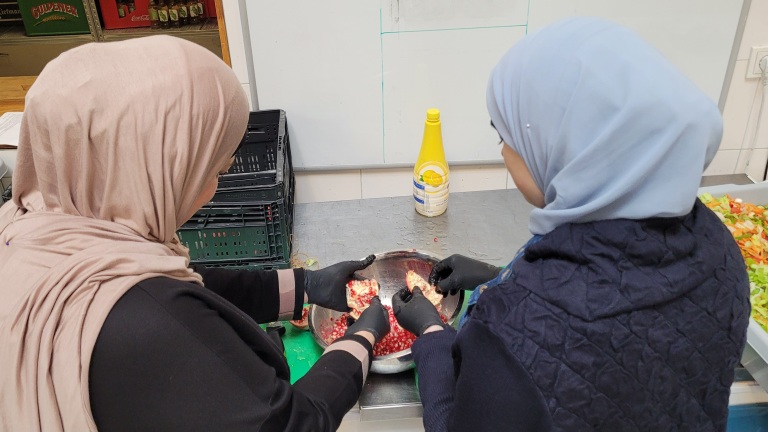 Twee vrouwen met hoofddoek en handschoenen maken een granaatappel schoon boven een schaal in een keuken