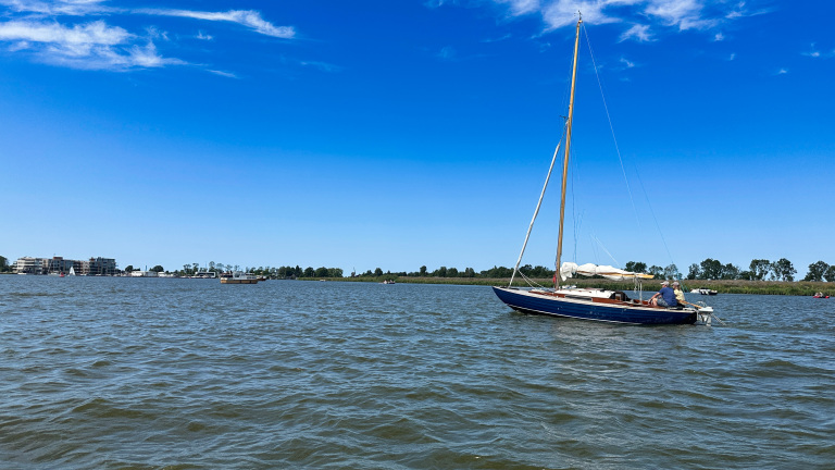 Blauwe zeilboot met gestreken zeil op een meer, met twee mensen aan boord en een heldere blauwe lucht