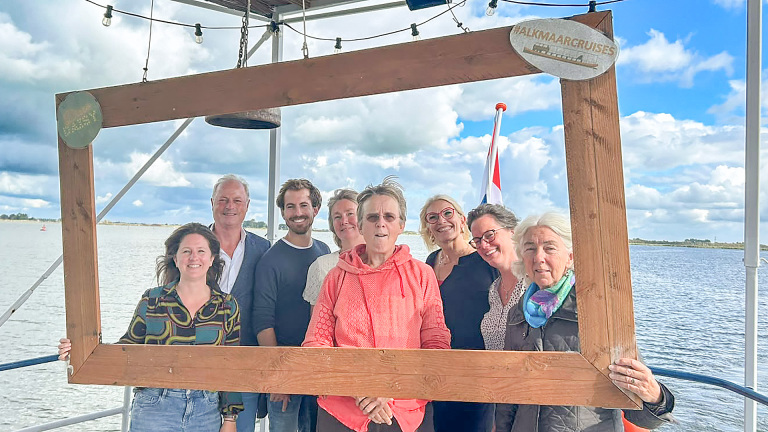 Groep mensen op een boot poseert met een groot houten fotolijstkader; op het bord staat “#ALKMAARCRUISES”