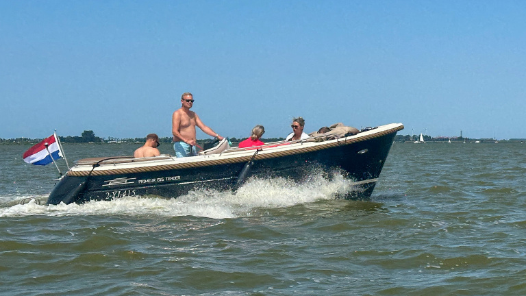 Open motorboot met Nederlandse vlag en vier opvarenden vaart over het water, met opspattende golven onder blauwe lucht