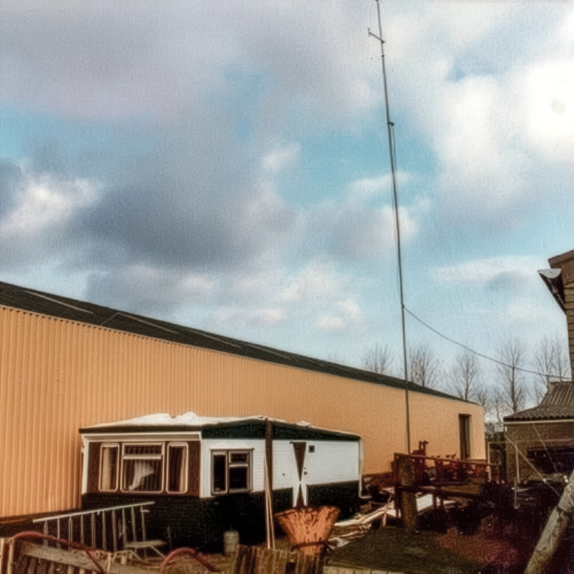 Hoge antennemast naast een loods en woonhuisjes op een erf, met bewolkte lucht op de achtergrond