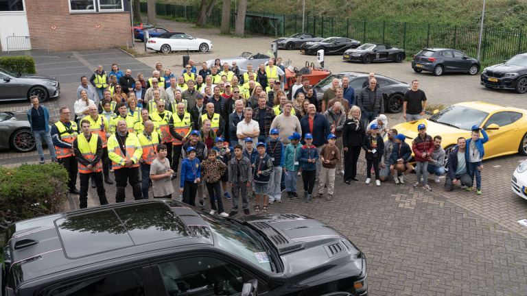 Grote groepsfoto op parkeerplaats met vrijwilligers in hesjes en kinderen met petten, omringd door sportauto’s