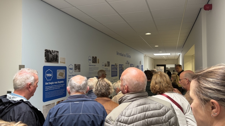 Groep bezoekers in een gang die informatiepanelen en foto’s op de muur bekijkt met tijdlijn “Het verhaal van Kaptein”