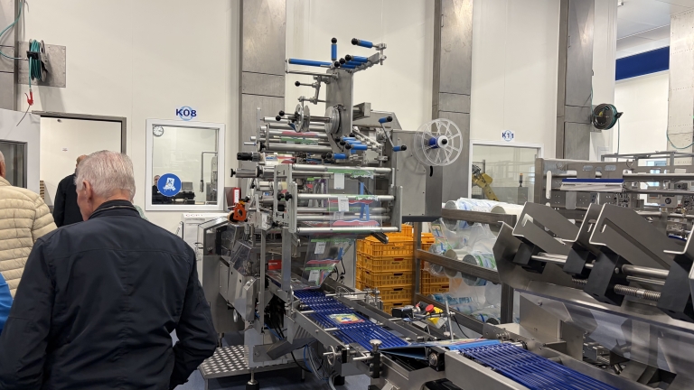 Verpakkingsmachine in fabriekshal met lopende band en plastic folierollen, terwijl bezoekers de productielijn bekijken