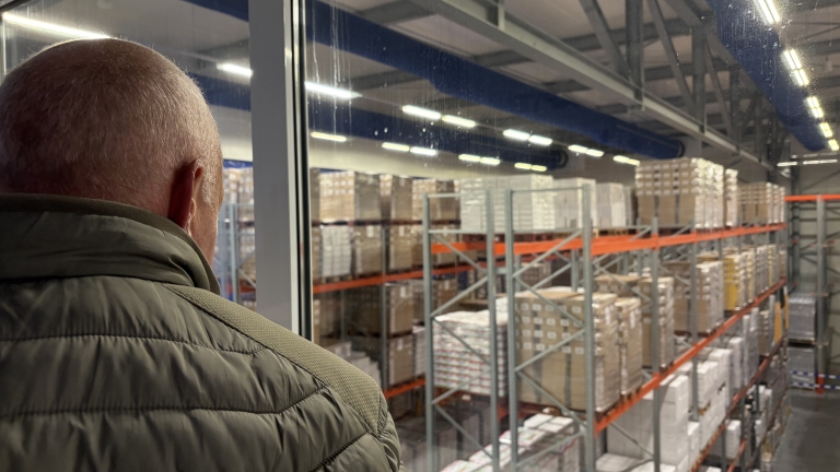 Man met kort haar kijkt door een raam uit over magazijnstellingen vol pallets en dozen in een distributiecentrum