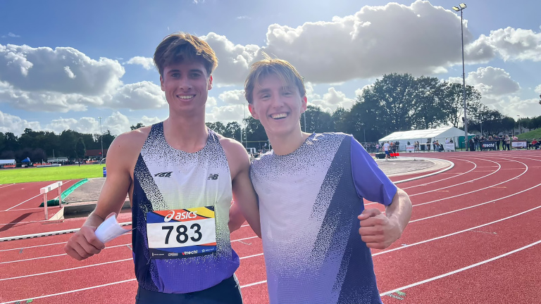 Twee lachende atleten op een atletiekbaan, één met startnummer 783, met bocht van de baan en wolkenlucht op de achtergrond