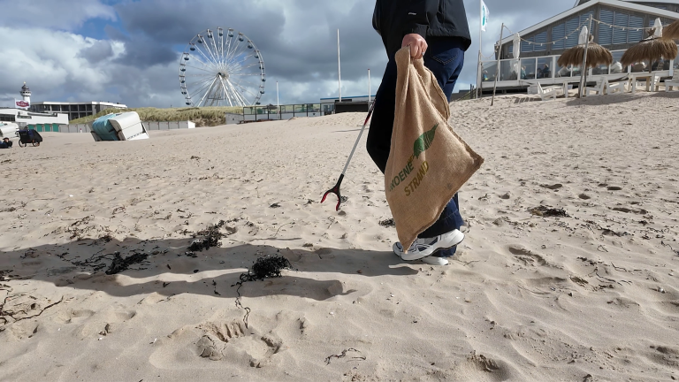 Persoon raapt afval op het strand met een grijper en jutezak; op de achtergrond een reuzenrad en strandpaviljoen