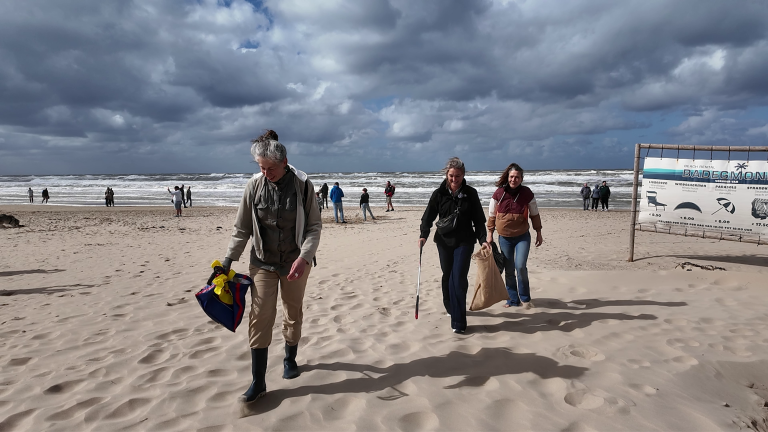 Drie vrouwen lopen over een winderig strand, één met tas, één met prikstok en één met jutezak, met zee op achtergrond