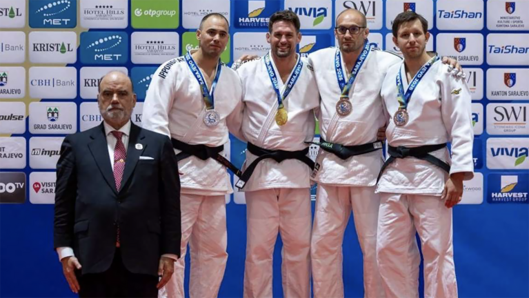 Vier judoka’s in witte gi met medailles poseren op het podium voor een sponsorwand, met een official in pak ervoor
