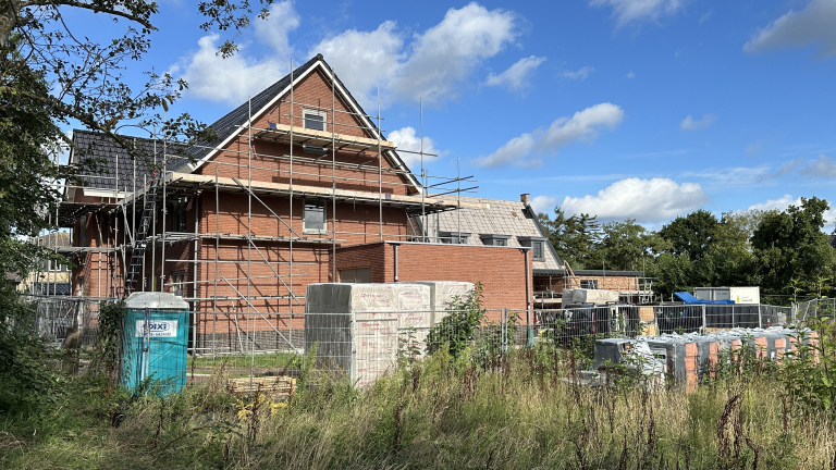 Bakstenen huis in aanbouw met steigers, bouwhekken en stapels bouwmateriaal onder een blauwe lucht