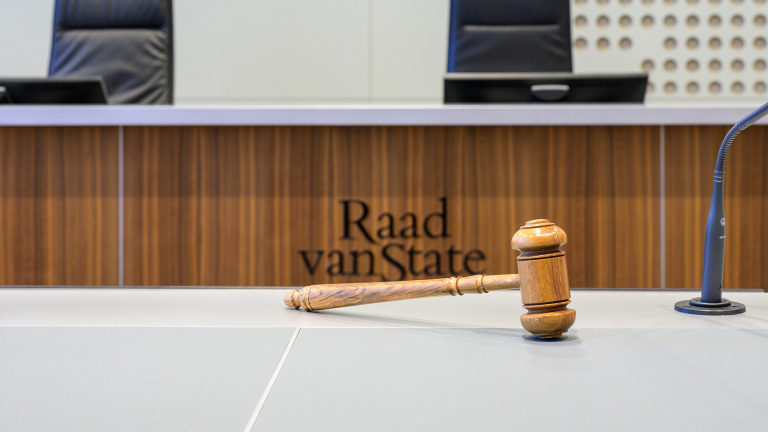 Houten hamer op tafel in rechtszaal, met op de achtergrond de tekst Raad van State en een microfoon