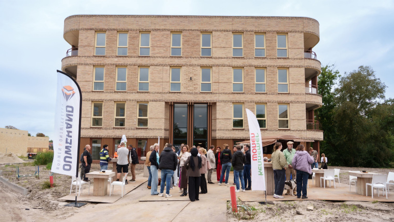 Groep mensen bij opening van nieuw bakstenen gebouw, met vlaggen “Oomen” en “reule deuel” voor de ingang