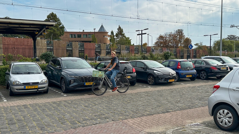 Man op fiets met groene krat rijdt langs geparkeerde auto’s bij een station, met bovenleiding en hek op de achtergrond