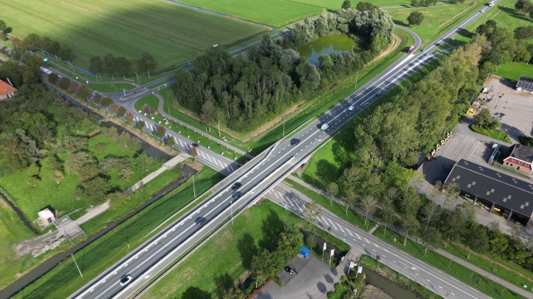 Luchtfoto van een snelwegviaduct over een kruising en kanaal, met auto’s, bomen, weilanden en een bedrijfsterrein