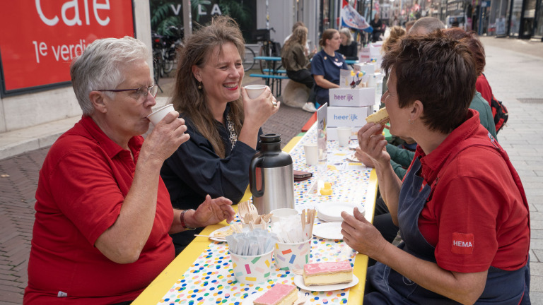 Drie mensen drinken koffie en eten gebak aan een lange tafel op straat tijdens een gezellig evenement