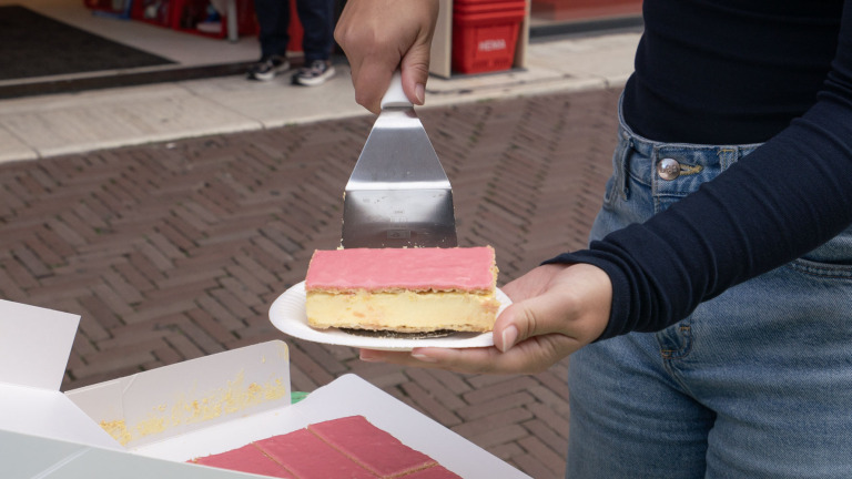 Hand met taartschep die een tompouce op een papieren bordje vasthoudt, met open doos tompoucen op de voorgrond