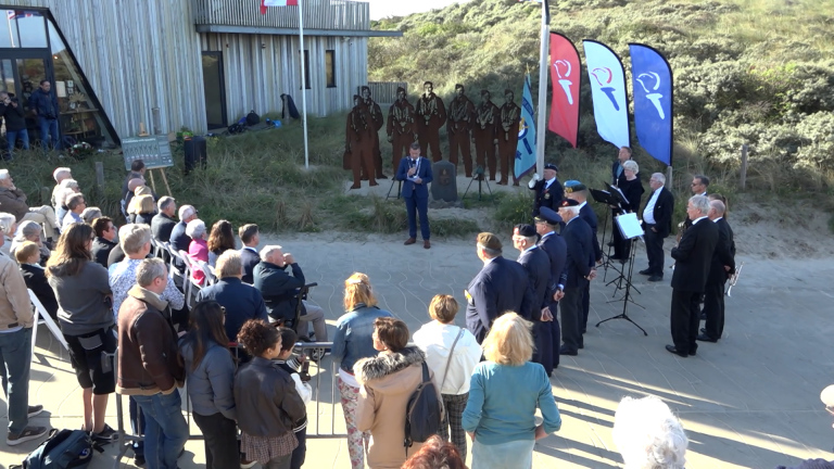 Bijeenkomst bij een monument in de duinen, met spreker, publiek en fanfare naast vlaggen