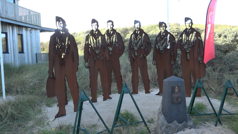 Stalen silhouetten van zes piloten in uniform bij een herdenkingsmonument met plaquette, naast duinen