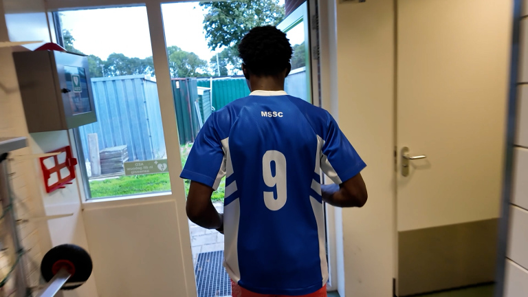 Persoon in blauw sportshirt met nummer 9 en tekst MSSC loopt door een open deur naar buiten
