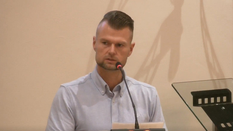 Man met opgeschoren kapsel spreekt in een microfoon tijdens een bijeenkomst, met glasplaat van lessenaar rechts