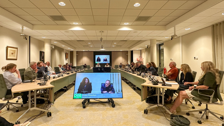Raadsvergadering in vergaderzaal met mensen rond U-vormige tafels en scherm met videobijdrage en naam “Peter Soonius”