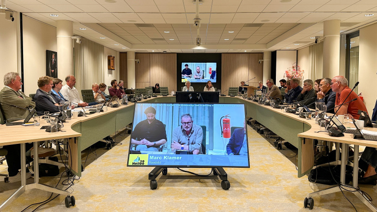 Raadsvergadering in vergaderzaal met raadsleden aan U‑vormige tafels en een scherm met live videobijdrage