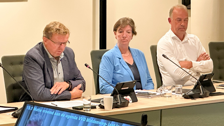Drie mensen zitten aan een vergadertafel met microfoons en tablets tijdens een raads- of commissievergadering