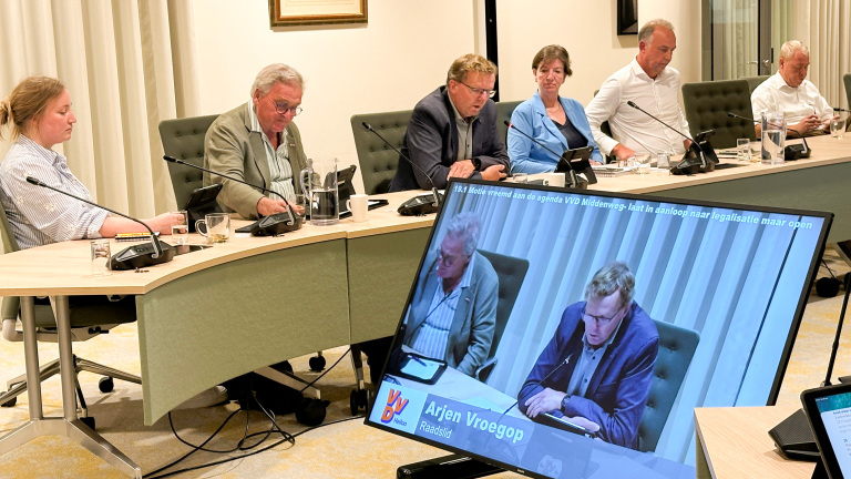 Raadsvergadering met zes mensen aan tafel met microfoons; op een groot scherm staat een spreker met tekst “Arjen Vroegop”
