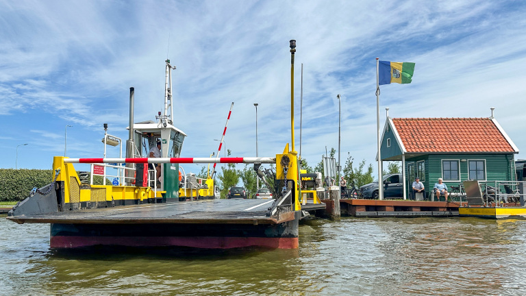 Pontveer met slagboom en oprit aan het water, met groen huisje en vlag op de kade onder blauwe lucht