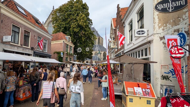 Drukke straatmarkt met kraampjes en bezoekers tussen historische panden, met rood-witte vlaggen en kerk op de achtergrond