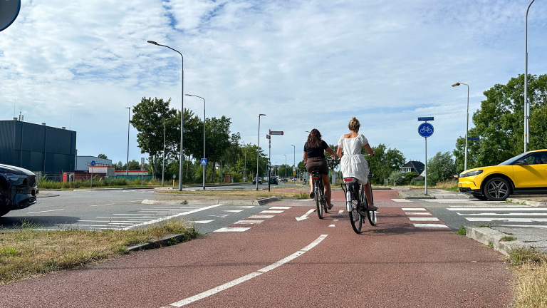 Twee fietsers rijden over een rood fietspad een kruising op, met auto's en verkeersborden op de achtergrond