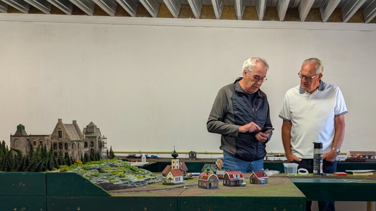 Twee mannen bekijken een smartphone boven een modelspoorbaan met miniatuurgebouwen, bos en trein op de achtergrond