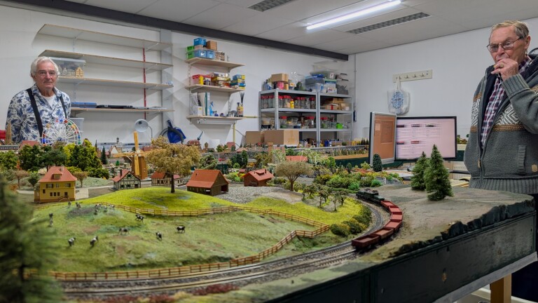 Twee mannen bekijken een modelspoorbaan met dorp, molen, bomen en trein in een werkplaats met planken en computer