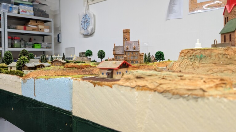 Modelspoorlandschap met kasteelachtig gebouw, huisjes en bomen op een heuvel, in een werkruimte met stellingen op de achtergrond
