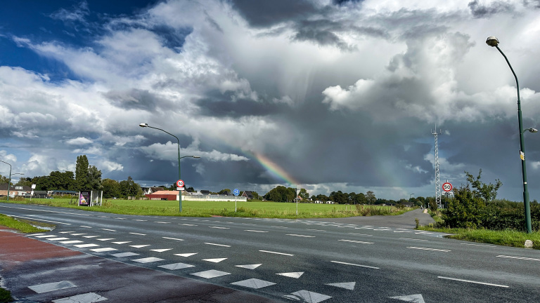 Regenboog boven groen veld en dorpje, met donkere onweerswolken en kruising met 50‑km-borden en lantaarnpalen