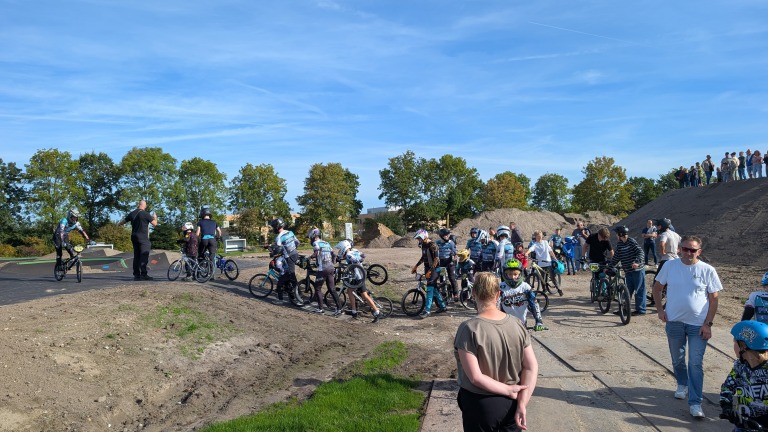 Groep BMX-rijders met helmen en fietsen staat klaar bij een baan, terwijl toeschouwers langs de rand kijken