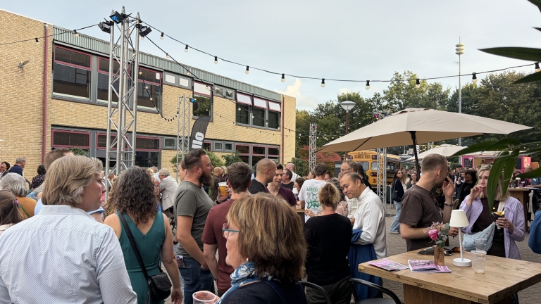 Drukbezocht buitenfeest op een plein met lichtslingers, mensen met drankjes en een gele schoolbus op de achtergrond