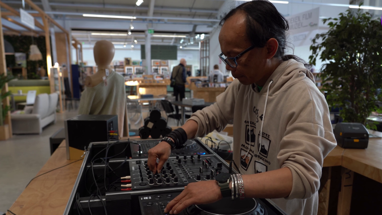 Persoon met bril bedient een DJ-mengpaneel op een tafel in een ruime winkel- of beursruimte