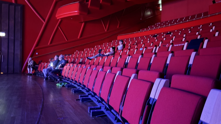 Rode theatertribune met bijna lege rijen stoelen; enkele toeschouwers zitten verspreid in de zaal