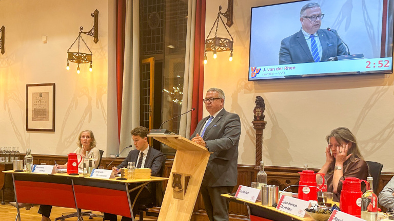 Man spreekt vanaf een katheder tijdens raadsvergadering; op scherm staat “J. van der Rhee” met aftellende spreektijd