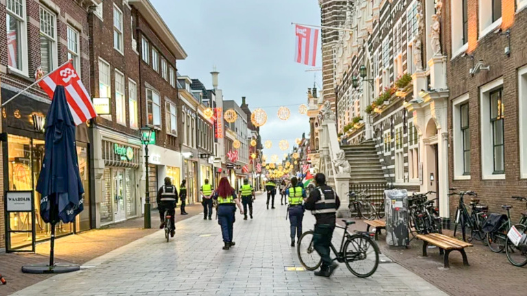 Winkelstraat met feestverlichting; agenten en handhavers lopen en fietsen langs winkels en gevels met vlaggen