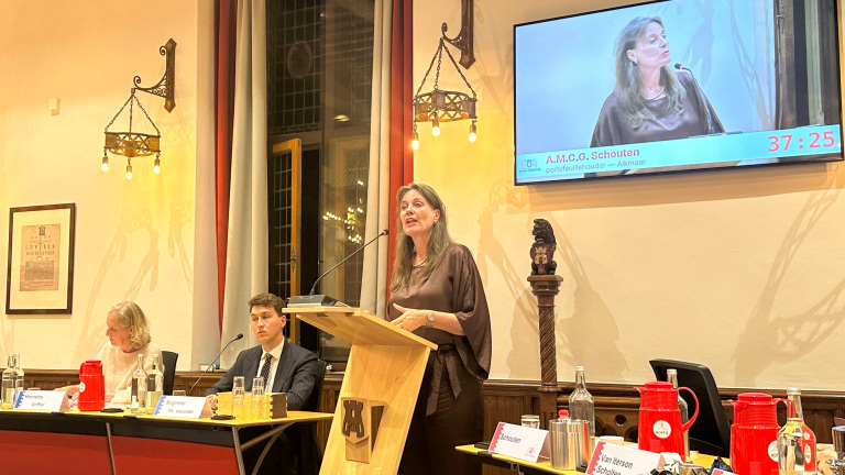 Vrouw spreekt achter een katheder in raadszaal; op scherm haar beeld met naam A.M.C.G. Schouten en timer, anderen zitten aan tafel