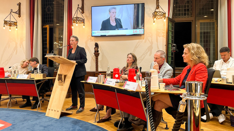 Vrouw spreekt achter een lessenaar tijdens raadsvergadering, met raadsleden aan tafels en haar beeld op een groot scherm